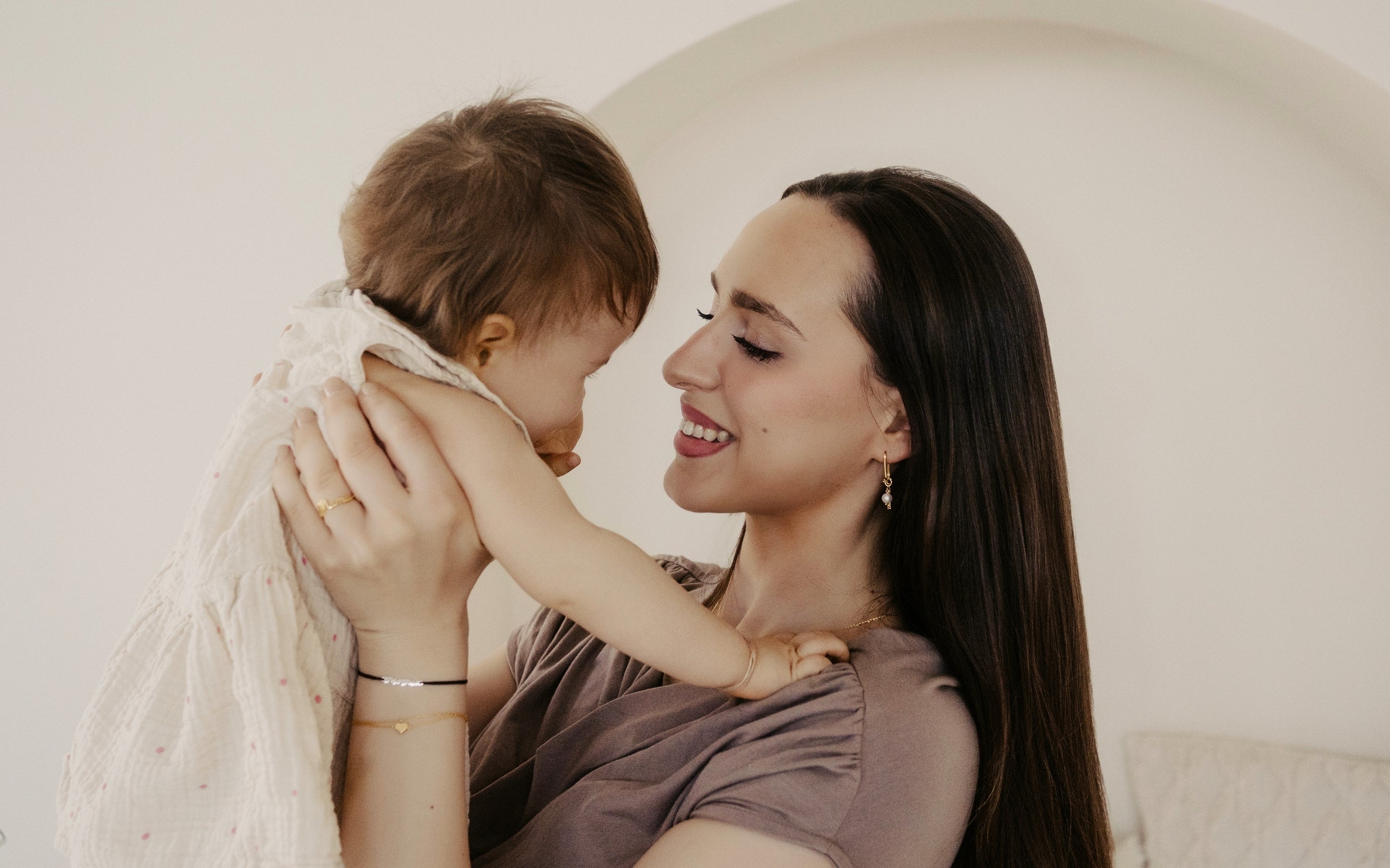 Nahaufnahme einer glücklichen Mutter, die ihr Baby liebevoll hochhält und anlächelt, während sie zarten Goldschmuck von Mama Collection trägt – ein Symbol für Mutterschaft, Nähe und besondere Geschenke für frischgebackene Mütter.
